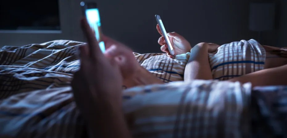 Duas pessoas deitadas na cama, de noite, antes de dormir, olhando seus celulares. 