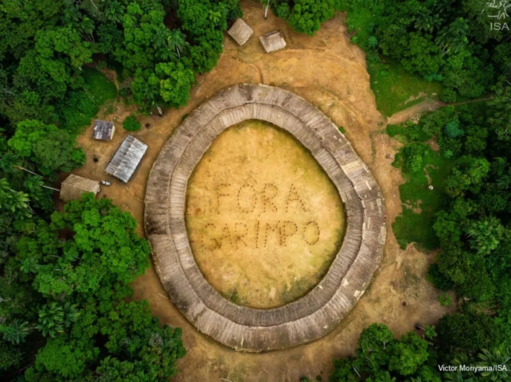 Imagem do centro de uma aldeia indígena onde se lê no chão: FORA GARIMPO