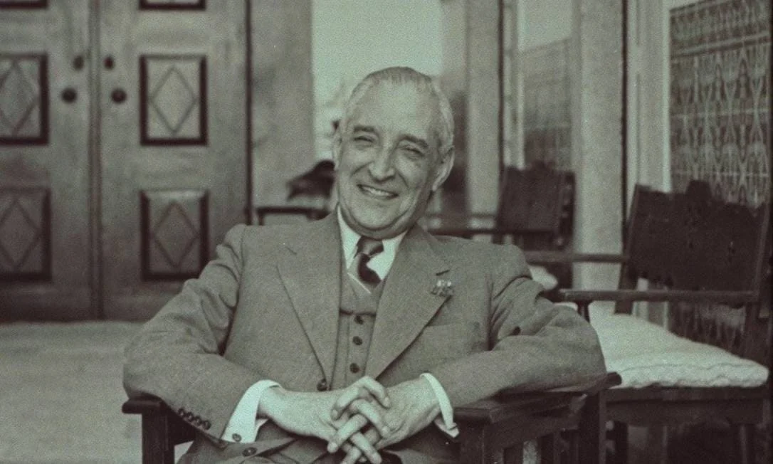 António de Oliveira Salazar sentado em uma cadeira olhando para frente, sorrindo
