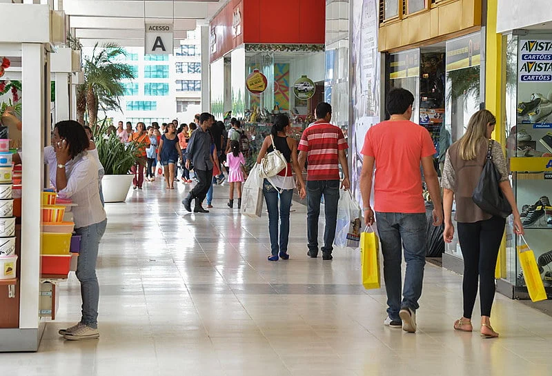 Interior de um shopping center movimentado, com várias pessoas andando e fazendo compras. Há lojas nas laterais e algumas barracas ou quiosques.