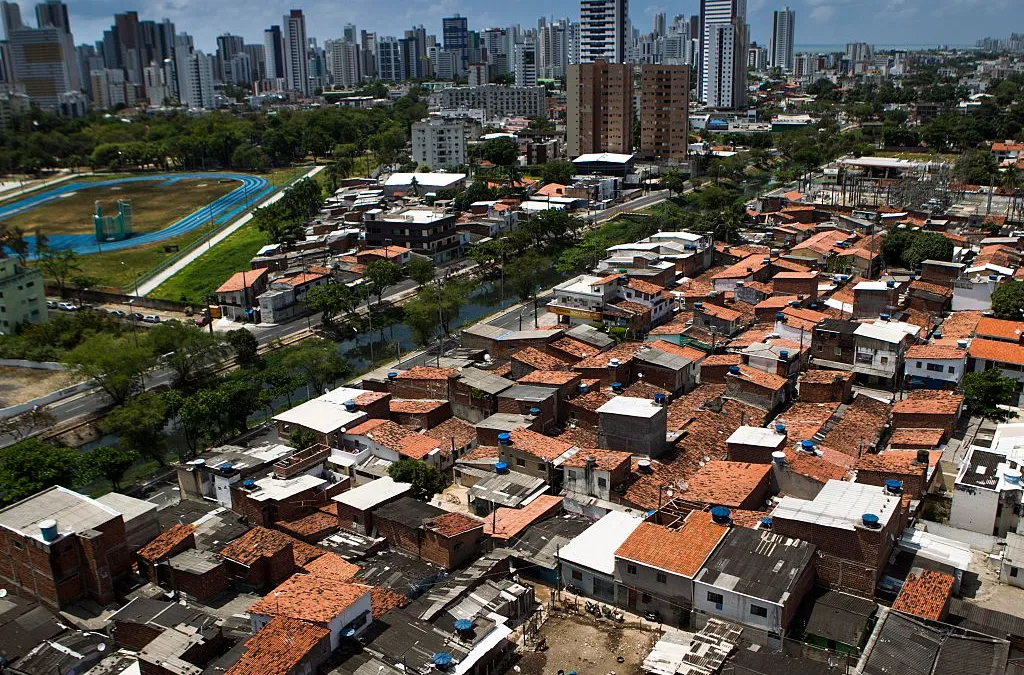 desigualdade social Paisagem urbana com um contraste marcante em Boa Viagem, no Recife, Brasil. Em primeiro plano, há uma área densamente povoada com muitas casas de telhados de barro e zinco, que se assemelham a uma comunidade mais humilde ou favela. Ao fundo, pode-se ver edifícios mais altos e modernos, que formam o horizonte de uma cidade. Há também um campo esportivo com uma pista azul visível à esquerda, e algumas áreas verdes intercaladas.