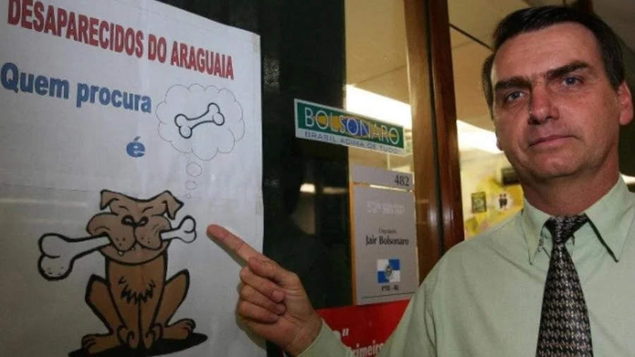 Em 2004, o então deputado Jair Bolsonaro posou em frente a um cartaz ironizando a busca pelos corpos dos desaparecidos da ditadura. Em 2023, já como Presidente, Bolsonaro cortou verbas para análise das ossadas encontradas no Cemitério de Perus, usado pelos agentes da repressão. Foto: reprodução