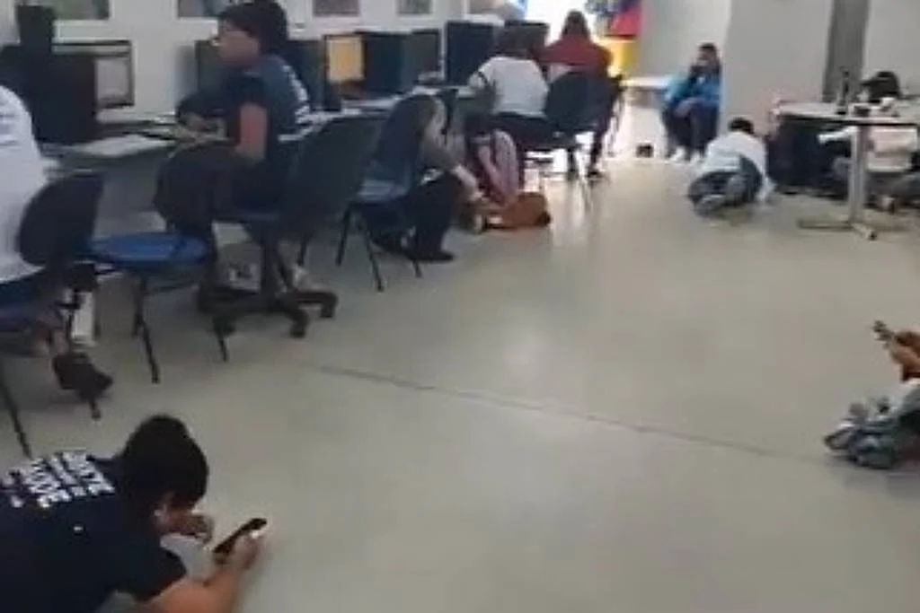Sala de aula ampla com janelas à esquerda. Alunos sentados em cadeiras com mesas, alguns usando máscaras. Professor em pé próximo à parede branca, interagindo com a turma. Piso claro e teto com luminárias embutidas.Funcionários se abrigam para escapar de tiros em unidade de saúde no Rio de Janeiro - Reprodução/Daniel Soranz no X