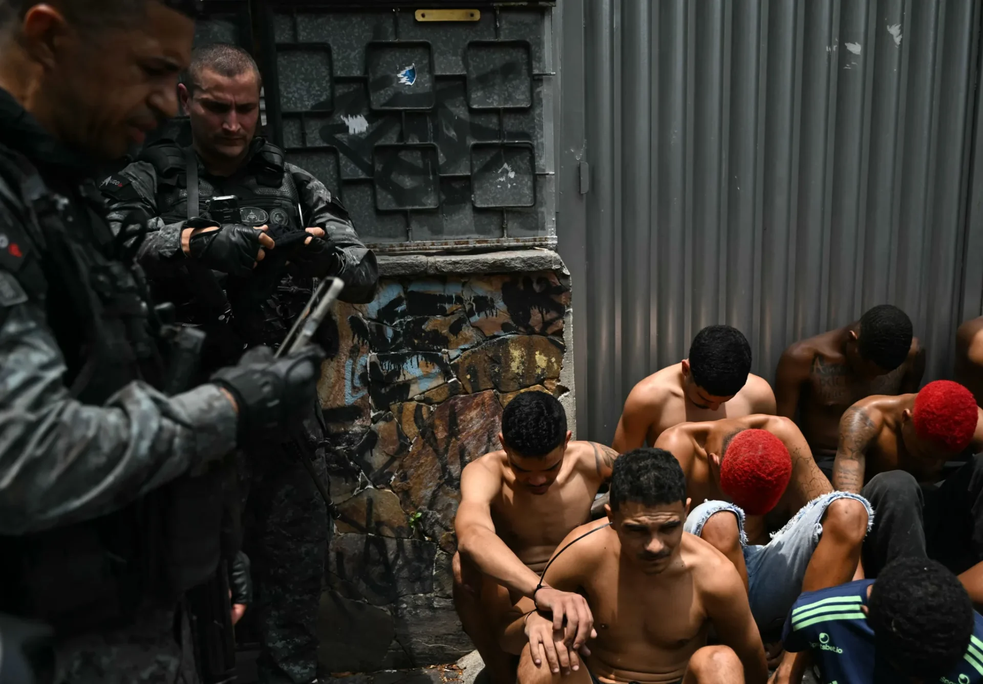 Um policial checa seu celular ao lado de suspeitos presos durante a Operação Contenção, no Complexo da Penha. | Crédito: Mauro Pimentel/AFP