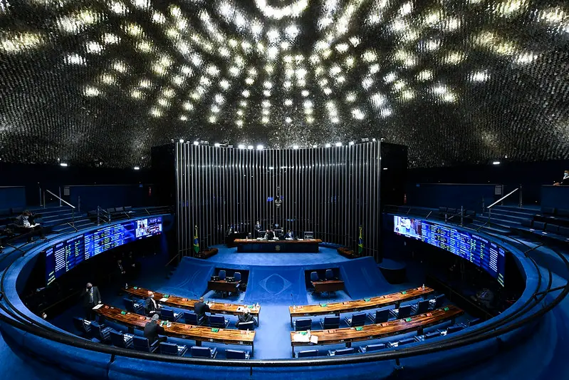 Ampla visão do Plenário do Senado Federal com parlamentares em seus assentos sob uma iluminação circular pontilhada no teto. Este ambiente representa o espaço de negociação política onde governo e legislativo decidem a alocação de recursos e a execução de emendas parlamentares