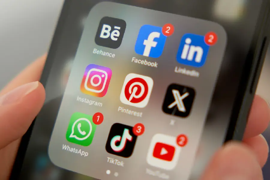 Fotografia em detalhe de uma mão segurando um celular que exibe aplicativos como Facebook, Instagram, TikTok, WhatsApp e YouTube, todos com notificações pendentes. No contexto do artigo, a imagem ilustra como os algoritmos dessas plataformas organizam a circulação de conteúdos, podendo amplificar o discurso de ódio ao priorizar publicações que geram alto engajamento por meio do medo ou da indignação.