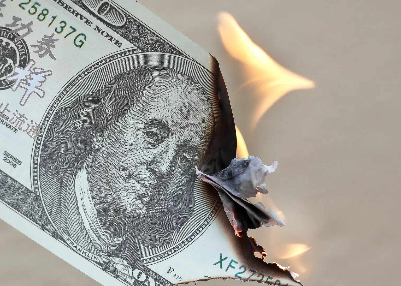 Grande plano de uma nota de 100 dólares americanos sendo consumida pelo fogo em uma das extremidades. A imagem simboliza os debates sobre desdolarização e a perda de hegemonia financeira dos Estados Unidos.
