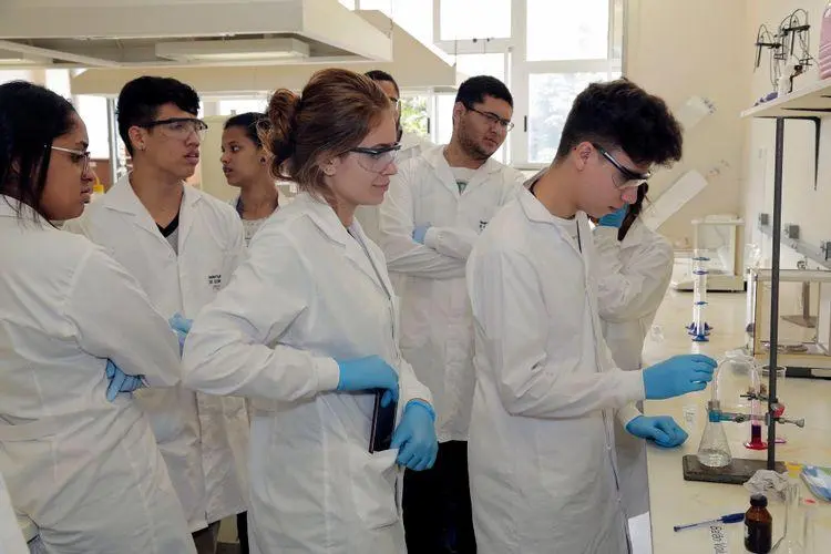 Jovens pesquisadores de jaleco branco operando equipamentos em um laboratório universitário da UFMG. O elemento visual conecta-se ao aumento expressivo de repasses via emendas para universidades federais, que buscam compensar cortes orçamentários e garantir a manutenção de projetos de pesquisa e infraestrutura acadêmica.