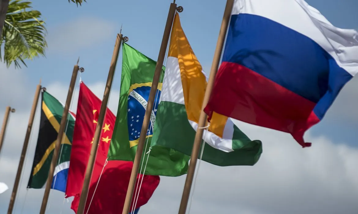 Fileira de bandeiras das nações do BRICS, incluindo África do Sul, China, Brasil, Índia e Rússia, ondulando ao vento sob um céu azul. A imagem ilustra a cooperação diplomática entre as potências emergentes do Sul Global.