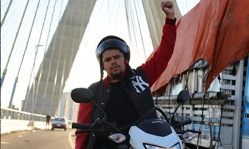 Paulo "Galo", ativista dos entregadores, posa sobre uma motocicleta com o punho erguido em sinal de luta, tendo uma ponte estaiada ao fundo. A imagem simboliza o desafio da organização coletiva em um modelo de trabalho individualizado e a importância da soberania digital para enfrentar o domínio das grandes plataformas.