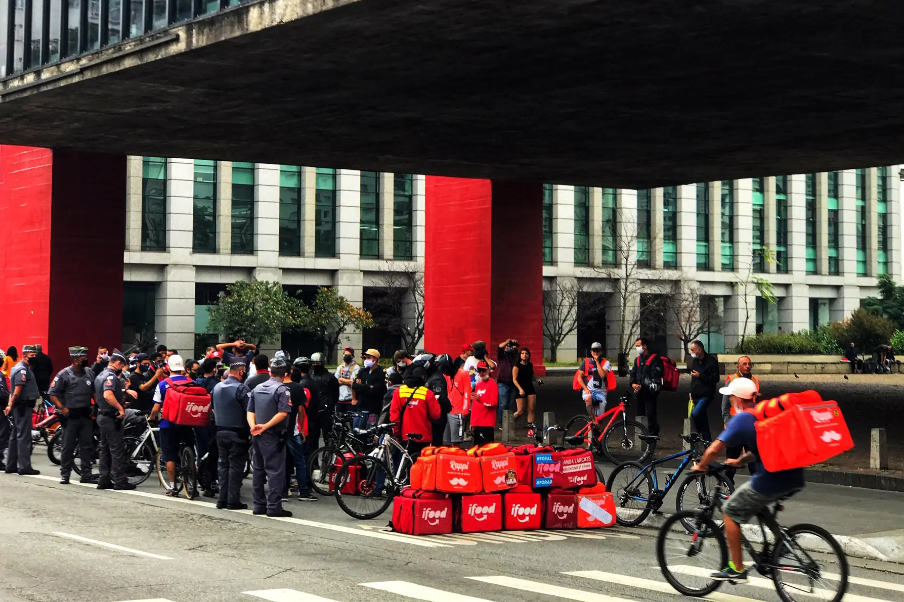 Grupo de entregadores de aplicativos, com suas mochilas térmicas vermelhas e bicicletas, reunidos em um ato por melhores condições de trabalho em uma avenida urbana. O registro destaca a resistência contra a gestão por algoritmos e a busca por direitos básicos diante de jornadas extensas e baixa proteção social.
