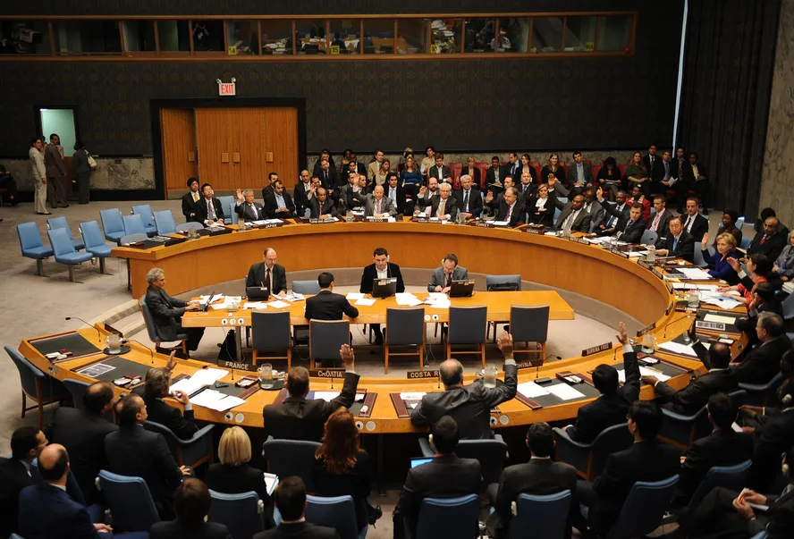 Vista aérea da mesa circular em formato de ferradura do Conselho de Segurança da ONU em Nova York, com delegados sentados e alguns com mãos levantadas em votação. O elemento destaca o centro das decisões da governança mundial.