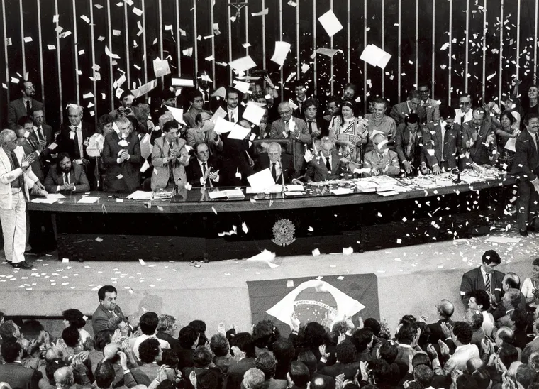 Sessão solene na Câmara dos Deputados Constituição de 1988 Foto em preto e branco retratando a sessão solene na Câmara dos Deputados no dia da promulgação da Constituição de 1988. Na imagem, parlamentares jogam papéis para o alto em comemoração. O documento pode ser considerado um marco na ampliação de mecanismos de participação social.