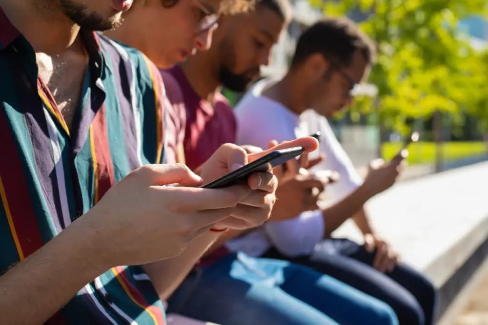 Um grupo de jovens sentados em um banco ao ar livre, cada um concentrado na tela de seu próprio celular. O registro visual destaca a mudança de hábito geracional e a queda no engajamento com mídias tradicionais, evidenciando que apenas 46% dos jovens de 16 a 24 anos consomem notícias de veículos jornalísticos diariamente.