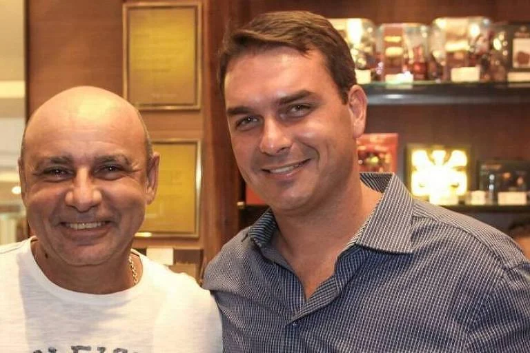 Fabrício Queiroz está à esquerda da foto. Ele é careca e veste camiseta branca. Está sorrindo. Flávio, do seu lado, é branco, tem cabelo castanho com um pequeno topete. Usa camisa em tom cinza e também sorri. Ao fundo, prateleiras com produtosFabrício Queiroz e Flávio Bolsonaro em foto antiga, de quando Flávio inaugurou loja de chocolates - Reprodução