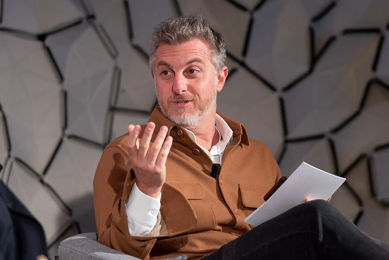 Fotografia do apresentador Luciano Huck gesticulando durante um painel no Fórum Econômico Mundial. A imagem ilustra a projeção institucional do apresentador, contrastando sua atuação como articulador social e palestrante com episódios de litígios jurídicos e ambientais de repercussão nacional, como o caso da ocupação de área costeira em Angra dos Reis.