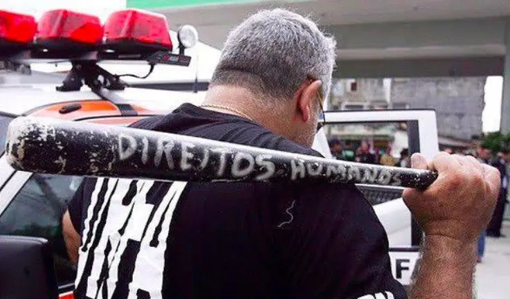Fotografia de uma pessoa segurando nas costas um bastão de madeira onde está escrito manualmente "Direitos Humanos". A imagem ilustra a deslegitimação do conceito no debate público brasileiro, onde o termo muitas vezes é associado à violência ou à ideia seletiva de "direitos humanos para humanos direitos".