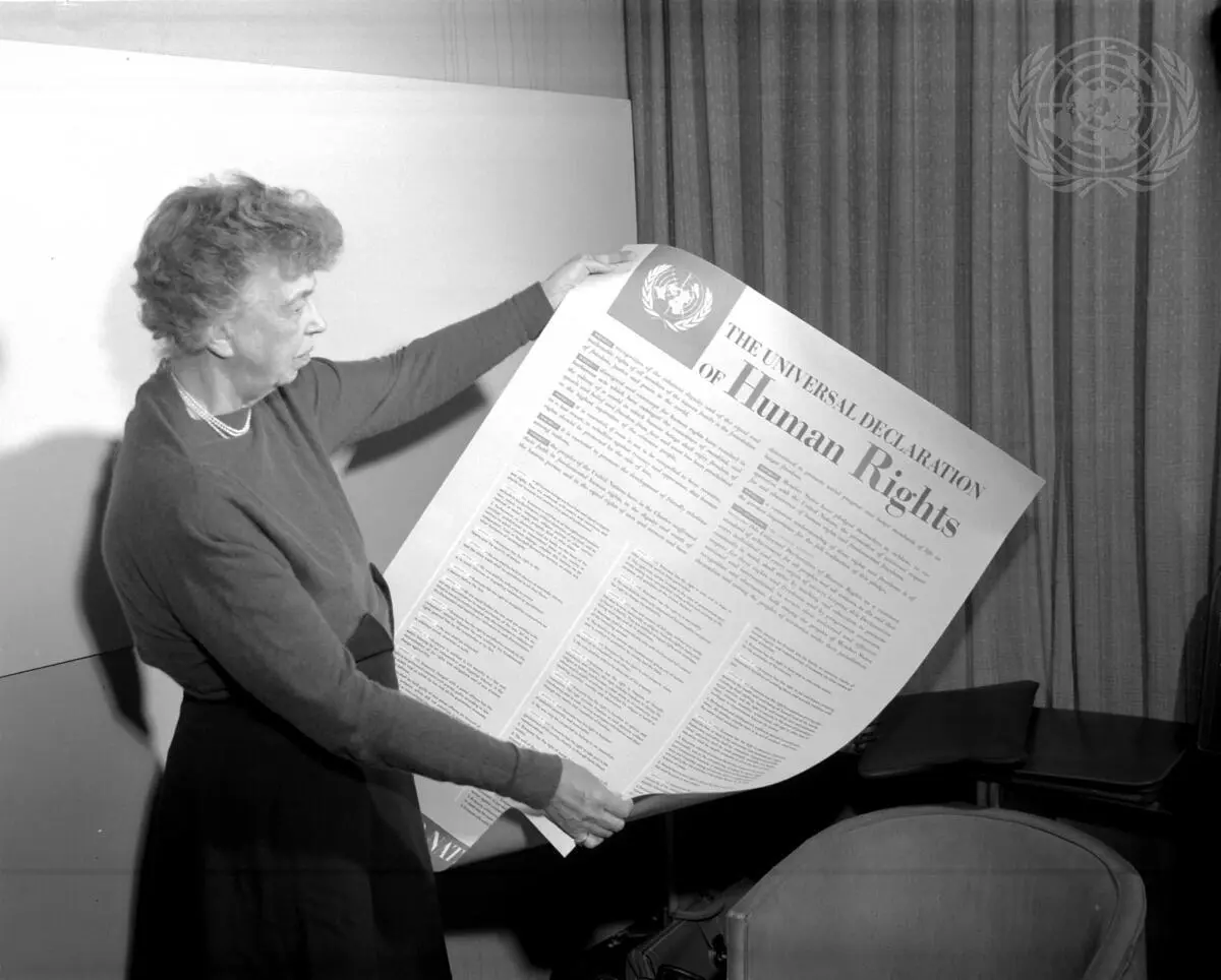 Fotografia em preto e branco de Eleanor Roosevelt segurando uma grande impressão da Declaração Universal dos Direitos Humanos. O registro marca a aprovação do documento após a Segunda Guerra Mundial como resposta aos horrores do Holocausto e para estabelecer padrões mínimos de proteção à vida.