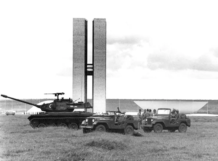 Fotografia histórica mostra tanques e jipes do Exército Brasileiro posicionados em frente às torres do Congresso Nacional em 1964. A imagem reforça a presença histórica dos militares na política brasileira, um elemento de influência que persistiu mesmo após a redemocratização devido a uma transição conduzida sem rupturas institucionais severas.