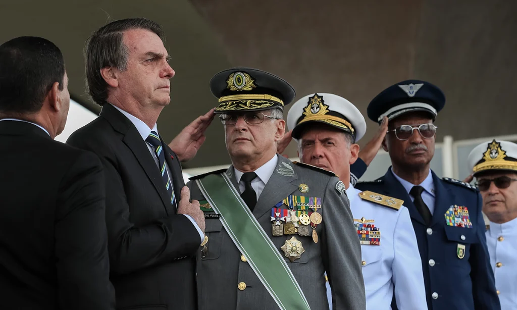 Ex-presidente Jair Bolsonaro de terno e gravata em posição de continência ao lado de militares fardados com condecorações, durante cerimônia oficial. A imagem acompanha a seção sobre autoritarismo e desinformação, ilustrando o vínculo simbólico entre lideranças políticas e instituições militares como elemento recorrente em contextos em que o medo e a construção de ameaças reorganizam o debate democrático.