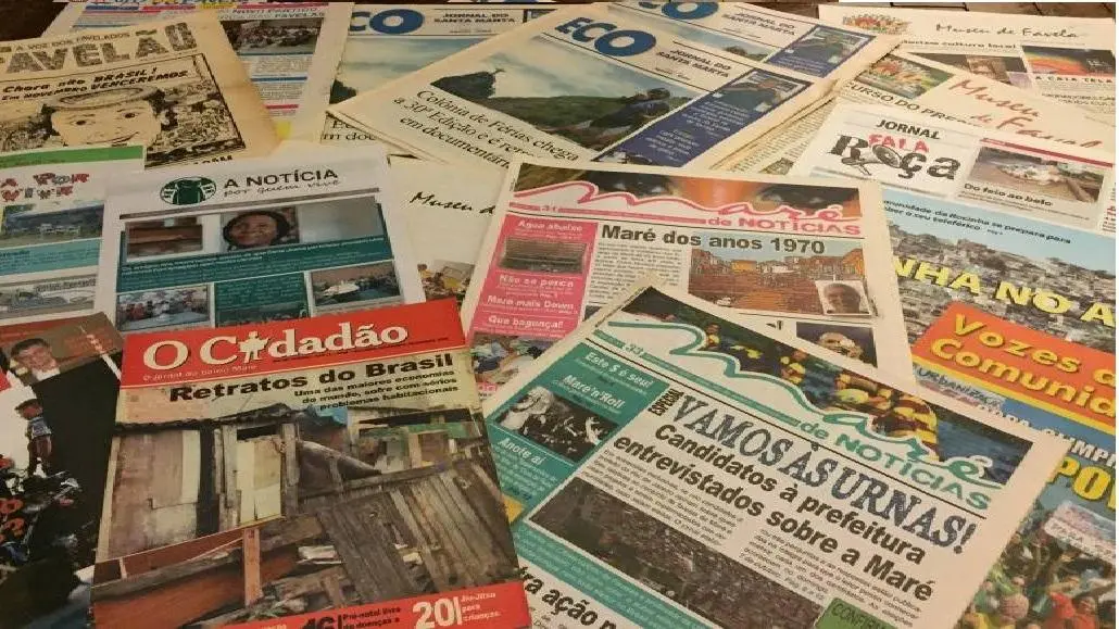 Diversos exemplares de jornais comunitários produzidos em favelas do Rio de Janeiro, com títulos como 'O Cidadão', 'Vozes da Comunidade' e 'Maré de Notícias', dispostos sobre uma superfície. A imagem representa o crescimento do jornalismo periférico no Brasil como contraponto à narrativa da mídia tradicional, em que moradores das periferias passam a disputar a própria representação, ampliando os temas abordados além do crime e da carência.