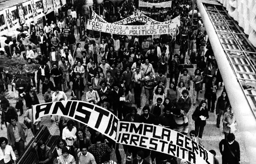 Imagem aérea em preto e branco de uma manifestação de rua onde se destaca uma grande faixa com os dizeres "Anistia Ampla, Geral e Irrestrita". A cena ilustra a mobilização social pela Lei da Anistia de 1979, marco que permitiu o retorno de exilados, mas que também garantiu a impunidade de agentes do Estado por crimes cometidos durante a repressão militar.