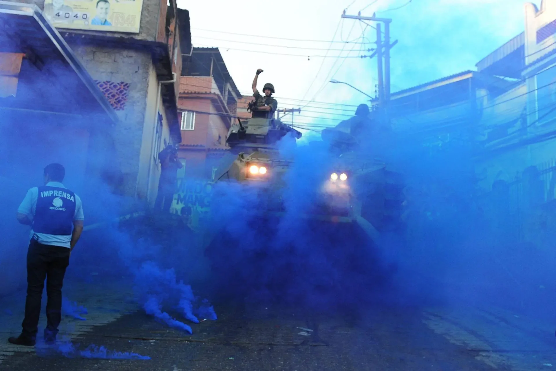Fotógrafo com colete de imprensa registra uma ação policial no Morro da Mangueira, em 2011, em meio a uma densa fumaça azul nas ruas da comunidade. A cena representa o padrão histórico de cobertura midiática das periferias brasileiras, em que a presença da imprensa nos territórios pobres se concentra em contextos de conflito e operações de segurança pública, contribuindo para associar pobreza à violência no imaginário social.