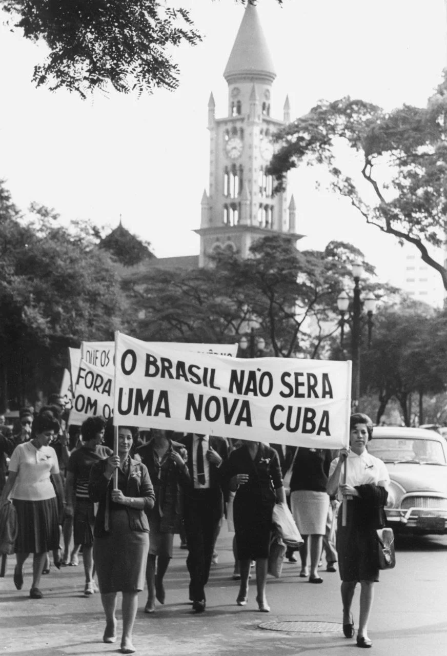 Foto histórica em preto e branco de mulheres em uma passeata segurando uma faixa com os dizeres "O Brasil não será uma nova Cuba". Ilustra o uso do anticomunismo para legitimar o golpe.