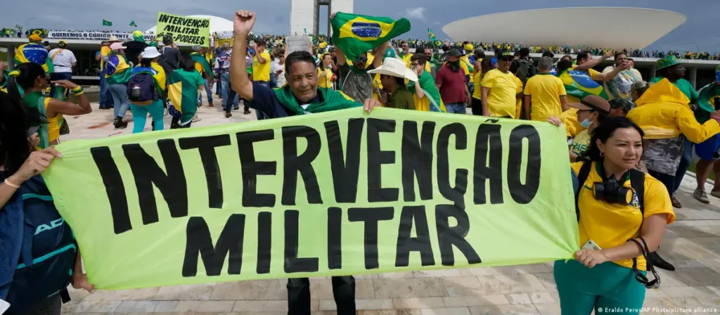 Manifestantes vestidos de verde e amarelo em Brasília segurando uma faixa verde neon com a frase "Intervenção Militar". A imagem conecta o saudosismo da ditadura aos ataques de 8 de janeiro.