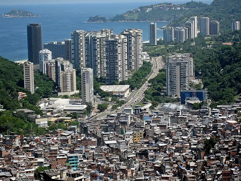Vista aérea da Favela da Rocinha, com suas construções densas e irregulares, em contraste direto com os edifícios residenciais de alto padrão do bairro de São Conrado, no Rio de Janeiro. A imagem ilustra visualmente a desigualdade social no Brasil, tema central do artigo, evidenciando como a separação geográfica entre ricos e pobres reflete estruturas históricas de exclusão que a mídia frequentemente reduz a dados econômicos.