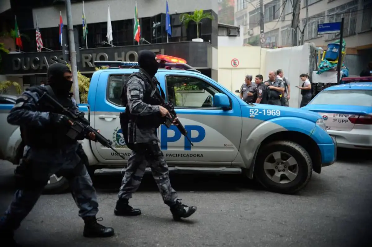 Dois policiais militares das Unidades de Polícia Pacificadora (UPP) fortemente armados e com o rosto encoberto caminham em posição de patrulha, com viaturas policiais ao fundo, nas comunidades do Cantagalo e Pavão-Pavãozinho, no Rio de Janeiro, em 2016. A imagem ilustra como a segurança pública se torna um dos principais gatilhos da política do medo: a ostensividade da presença policial em territórios periféricos reflete o cenário em que o debate eleitoral é deslocado para narrativas de ameaça e repressão, em detrimento de propostas estruturais.