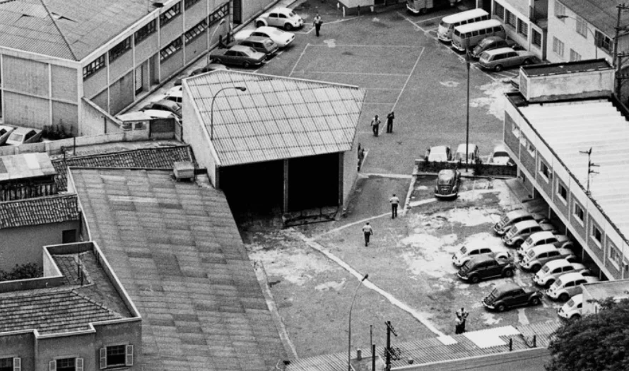 Fotografia aérea em preto e branco revela o pátio interno de um complexo de edifícios urbanos, com vários carros modelo Fusca estacionados e algumas figuras fardadas caminhando. Esta era a sede da Operação Bandeirantes (Oban) em São Paulo, local que se tornou o embrião do sistema DOI-CODI e simboliza a "profissionalização" da tortura na ditadura por meio da integração entre policiais e militares.