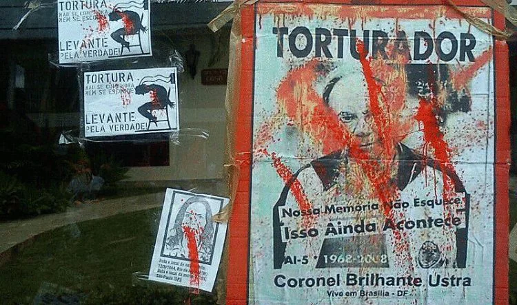 Fotografia de um "escracho" popular realizado em 2014 em frente à residência de Brilhante Ustra, com cartazes que o identificam como torturador. O elemento visual reforça a mobilização da sociedade civil contra a impunidade, destacando a importância da justiça de transição e do reconhecimento público dos crimes cometidos durante os anos de chumbo. Fotografia de um "escracho" popular realizado em 2014 em frente à residência de Brilhante Ustra, com cartazes que o identificam como torturador. O elemento visual reforça a mobilização da sociedade civil contra a impunidade, destacando a importância da justiça de transição e do reconhecimento público dos crimes cometidos durante os anos de chumbo.
