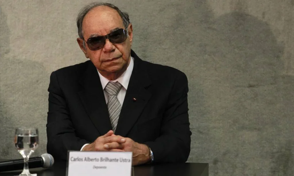 Fotografia do coronel reformado Carlos Alberto Brilhante Ustra durante seu depoimento à Comissão Nacional da Verdade em 2013. O registro captura o momento em que o primeiro oficial reconhecido pela Justiça como torturador confronta as investigações sobre crimes de Estado, ilustrando a tensão entre o revisionismo histórico e a busca por verdade e reparação. Fotografia do coronel reformado Carlos Alberto Brilhante Ustra durante seu depoimento à Comissão Nacional da Verdade em 2013. O registro captura o momento em que o primeiro oficial reconhecido pela Justiça como torturador confronta as investigações sobre crimes de Estado, ilustrando a tensão entre o revisionismo histórico e a busca por verdade e reparação.