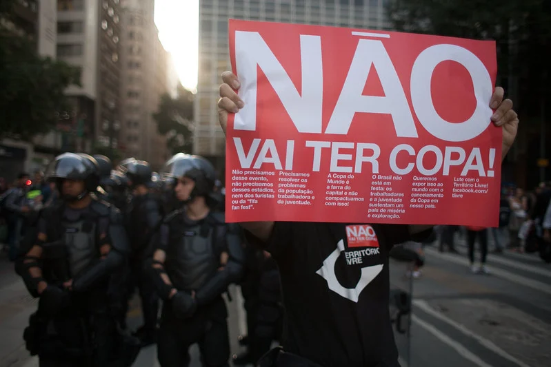Manifestante segurando cartaz vermelho com a frase "Não Vai Ter Copa" durante protesto nas ruas do Brasil em 2014, com policiais ao fundo