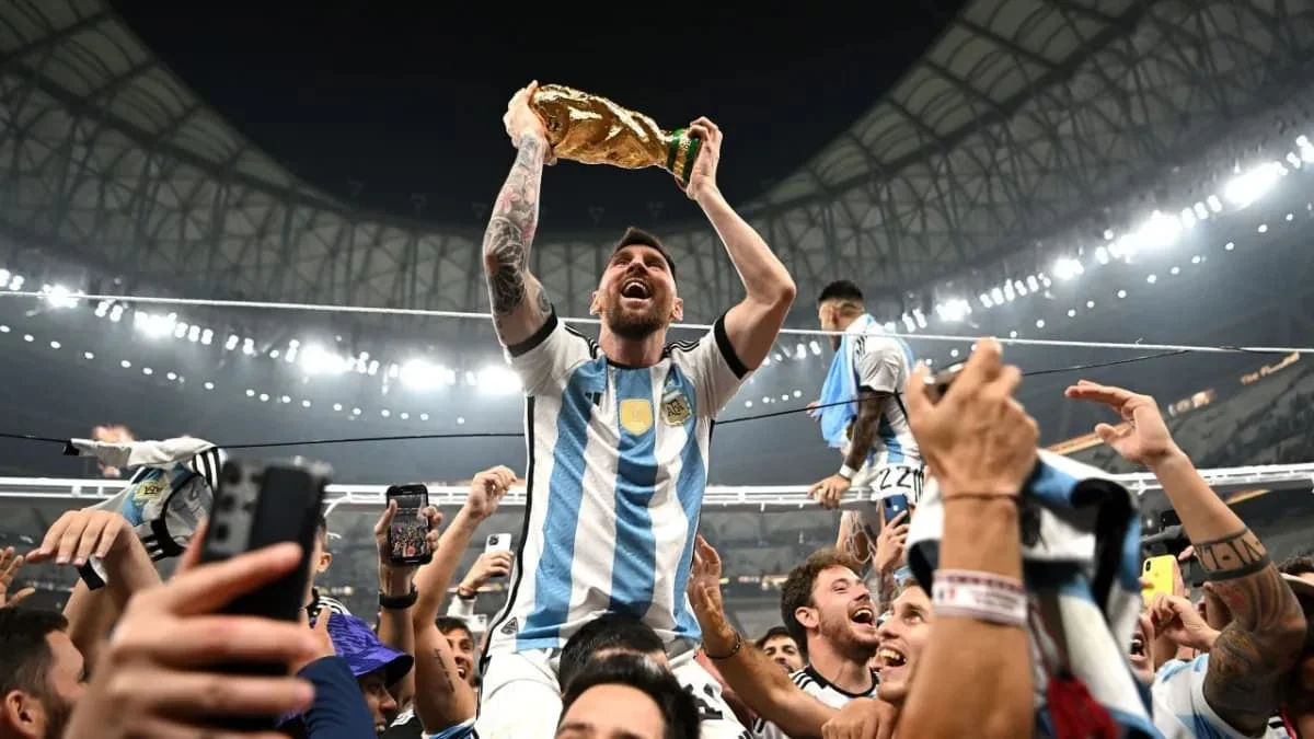 Lionel Messi erguendo a taça da Copa do Mundo de 2022 no Catar, com jogadores argentinos e torcedores ao fundo comemorando o tricampeonato