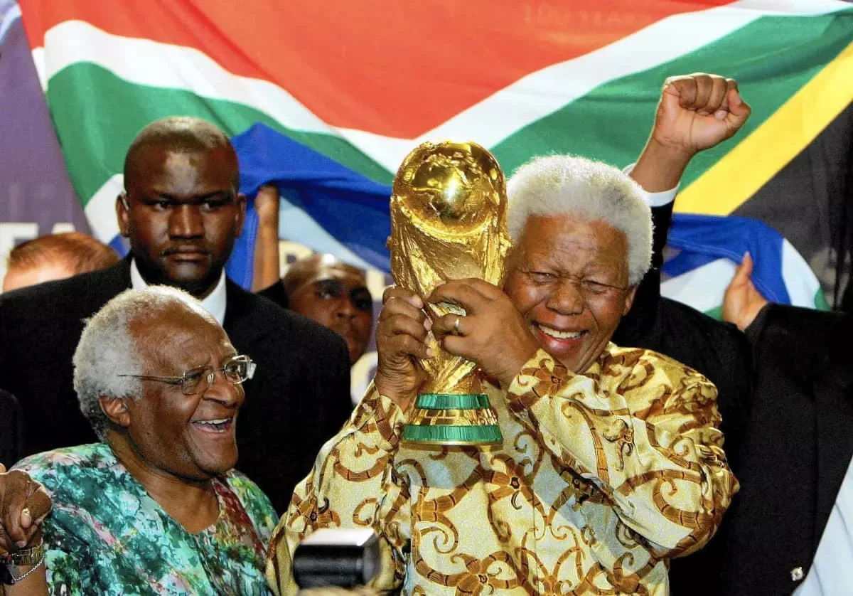 Nelson Mandela sorrindo e segurando a taça da Copa do Mundo de 2010 na África do Sul, com a bandeira sul-africana ao fundo