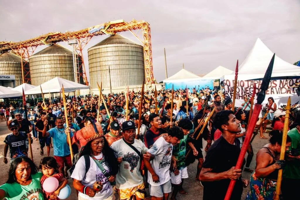 Povos indígenas marcham em frente às instalações da Cargill no Pará, em protesto contra a desestatização dos rios Madeira, Tocantins e Tapajós, carregando lanças e bandeiras em mobilização coletiva. A imagem evidencia como a disputa pelo orçamento público se conecta diretamente à disputa pelo território, com comunidades tradicionais reagindo ao avanço de grandes projetos econômicos sobre áreas próximas a seus territórios.