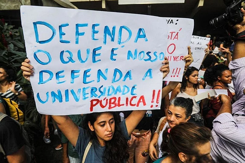 Manifestante segura cartaz com os dizeres 'Defenda o que é nosso — Defenda a Universidade Pública' durante protesto. A imagem representa a mobilização da comunidade acadêmica diante dos bloqueios e contingenciamentos orçamentários que afetaram universidades federais, institutos de pesquisa e agências de fomento científico no Brasil.