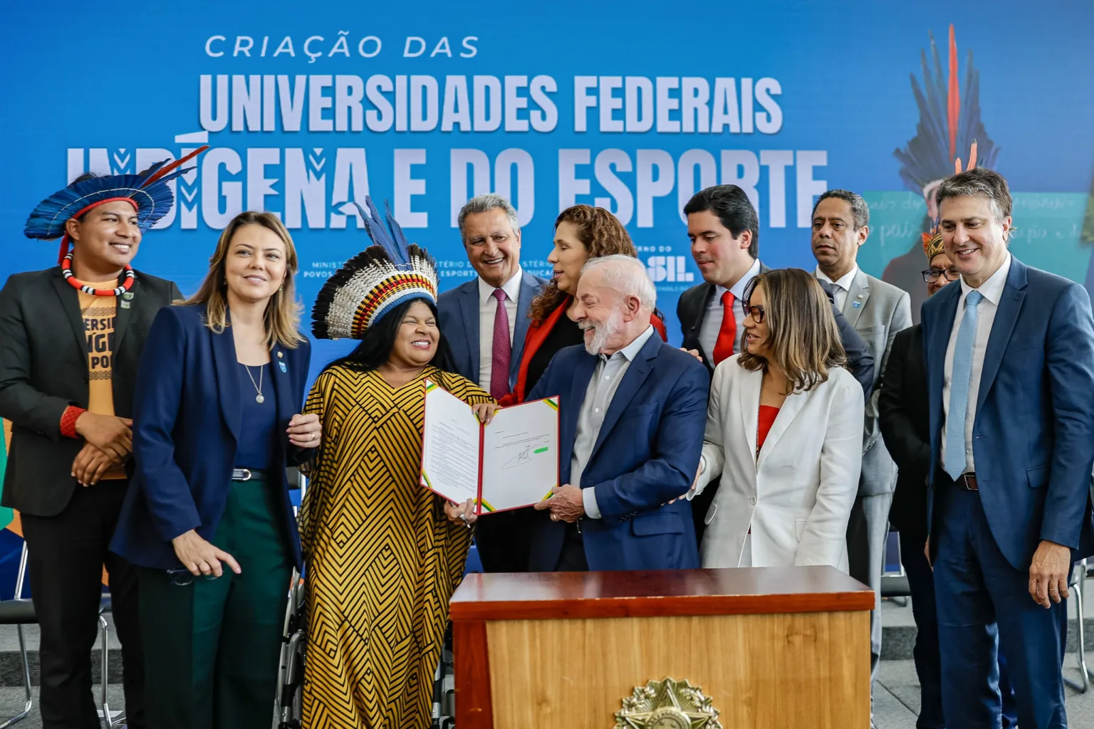 Presidente Lula assina documento em cerimônia oficial de criação das Universidades Federais Indígena e do Esporte, rodeado por lideranças indígenas e autoridades do governo. A imagem ilustra avanços institucionais na política educacional voltada a povos tradicionais, tema abordado na seção sobre o acesso desigual às políticas públicas para indígenas e quilombolas no Brasil.