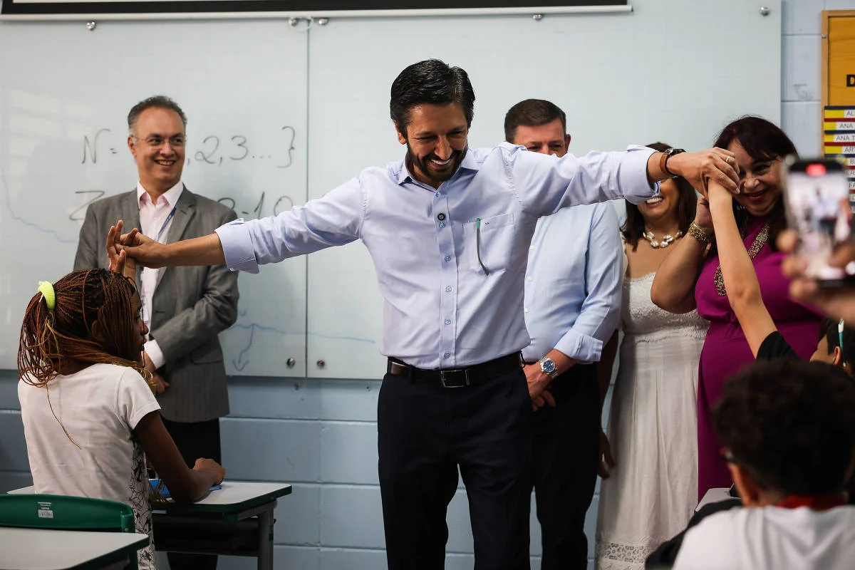 A imagem mostra um grupo de pessoas em uma sala de aula. Um homem de camisa clara está no centro, sorrindo e estendendo os braços, enquanto outras pessoas ao seu redor, incluindo uma mulher de vestido branco e outra de vestido rosa, parecem estar interagindo com ele. À frente, uma criança com cabelo trançado está sentada em uma mesa, observando a cena. Ao fundo, há uma lousa e outras pessoas assistindo. O prefeito Ricardo Nunes e o secretario de educação Fernando Padula em visita a escola municipal - Zanone Fraissat - 5.fev.25/Folhapress