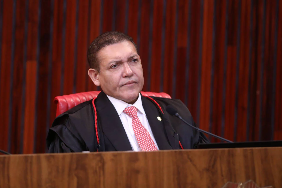 Homem de meia-idade veste toga preta com detalhes vermelhos e gravata vermelha com camisa branca, sentado atrás de bancada de madeira em ambiente com painéis verticais escuros ao fundo. O ministro Kassio Nunes Marques, em sessão do TSE - Luiz Roberto - 4.abr.26/TSE