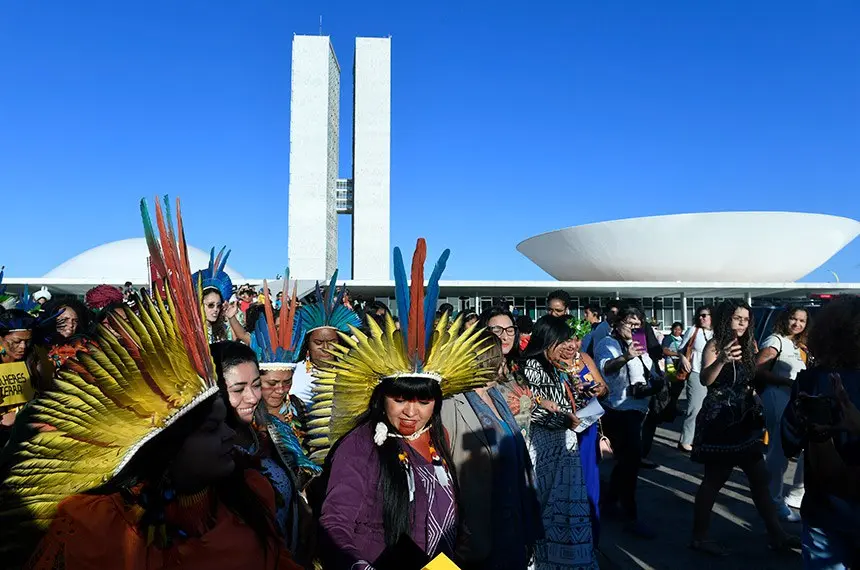 Indígenas usando cocares de penas coloridas participam de manifestação em frente ao Congresso Nacional, em Brasília, durante mobilização relacionada à defesa dos direitos territoriais dos povos originários.