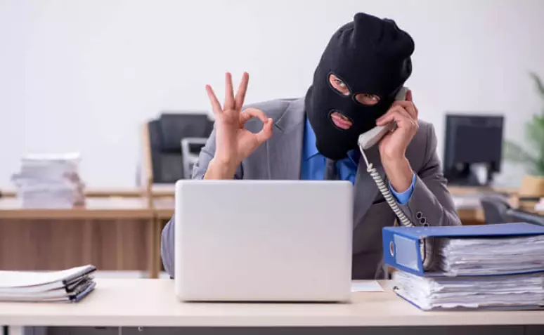 Homem usando balaclava preta, terno cinza e camisa azul fala ao telefone enquanto faz um gesto com a mão diante de um notebook aberto. Sobre a mesa há pilhas de documentos e pastas.