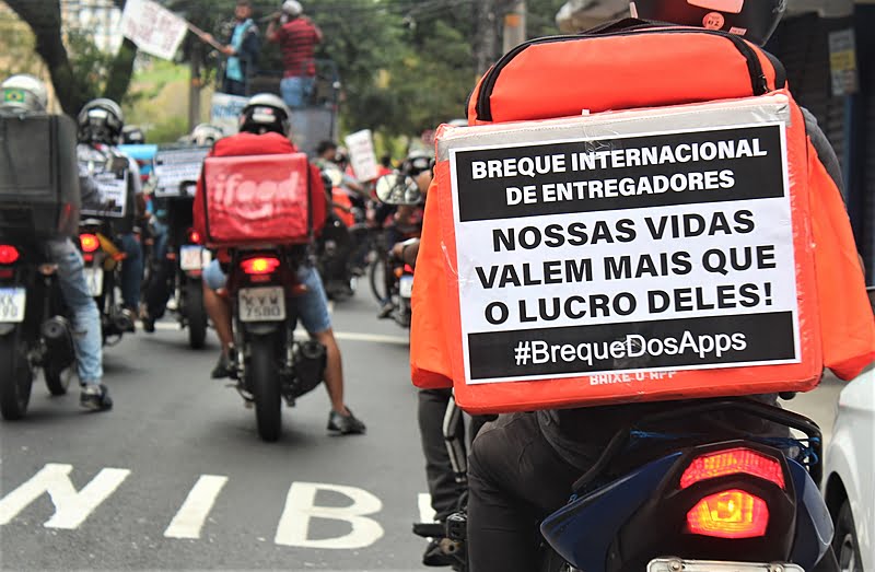 Fotogradia mostra paralisação feita por motociclistas entregadores por aplicativos. O foco está na traseira da mochila de um dos motociclistas, que possui um papel com os escritos: "Breque Internacional de entregadores. Nossas vidas valem mais que o lucro deles! #BrequeDosApps