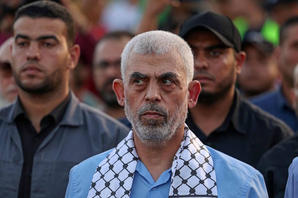 Um homem com cabelos grisalhos e barba, exibindo uma expressão séria e pensativa, está em primeiro plano. Ele veste uma camisa azul clara e um lenço palestino tradicional (keffiyeh) em volta do pescoço. Ao fundo, outros indivíduos desfocados observam atentamente, sugerindo que o homem pode estar participando de um evento ou manifestação.O chefe do braço político do grupo terrorista Hamas na Faixa de Gaza, Yahya Sinwar