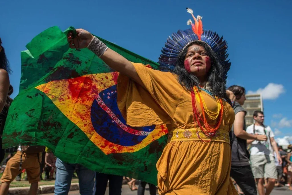 Indígena em manifestação com bandeira do Brasil ensanguentada. Foto: Divulgação. 